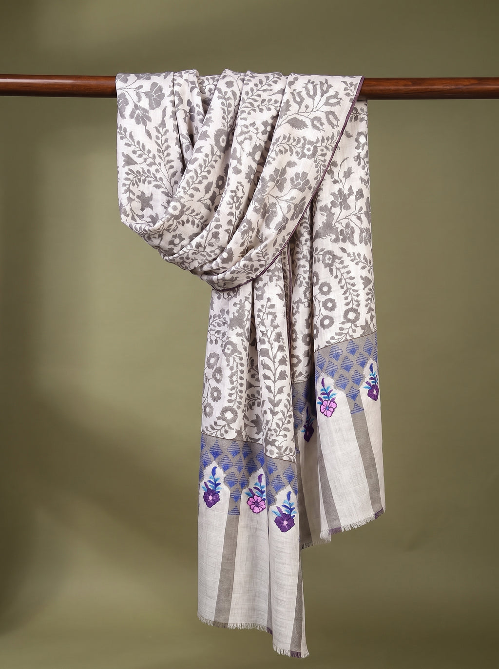 A light beige and white patterned shawl with purple floral accents near the border, draped over a wooden hanger against a solid olive green background.