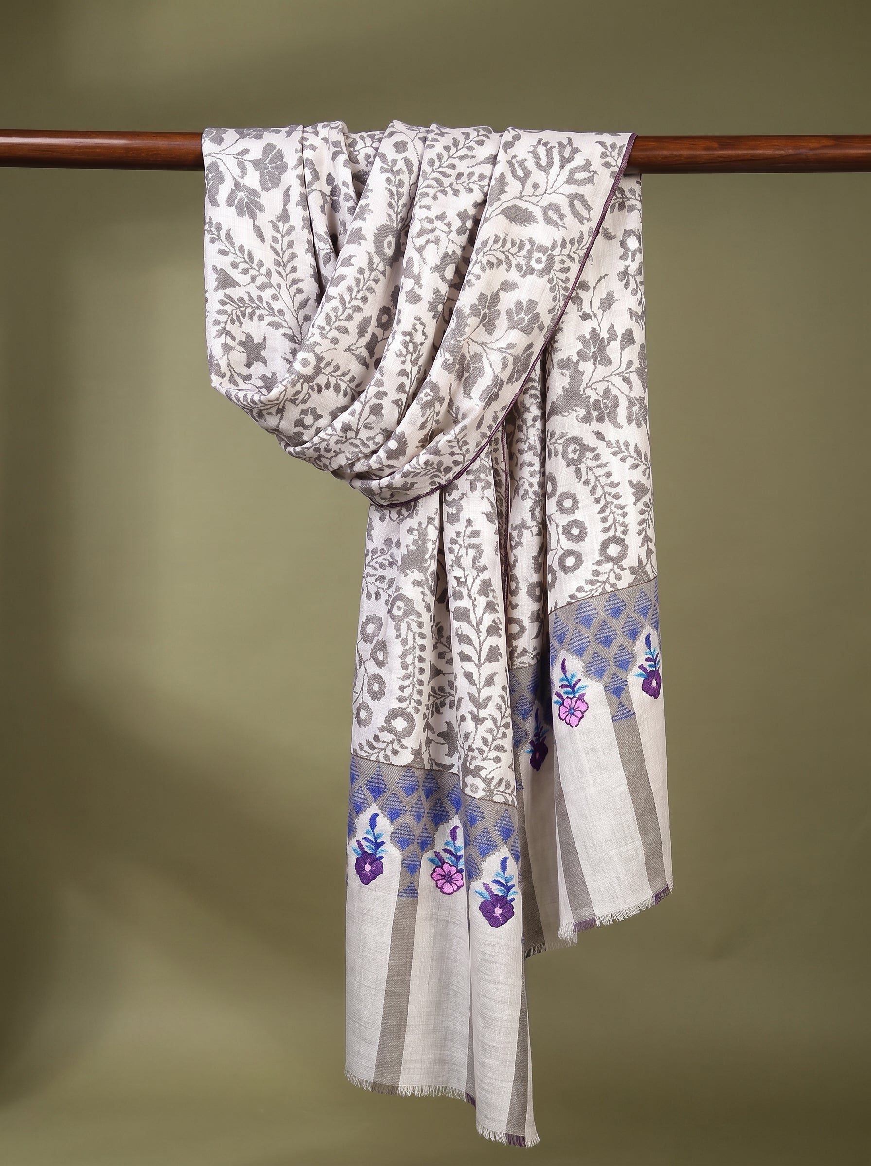 A light beige and white patterned shawl with purple floral accents near the border, draped over a wooden hanger against a solid olive green background.