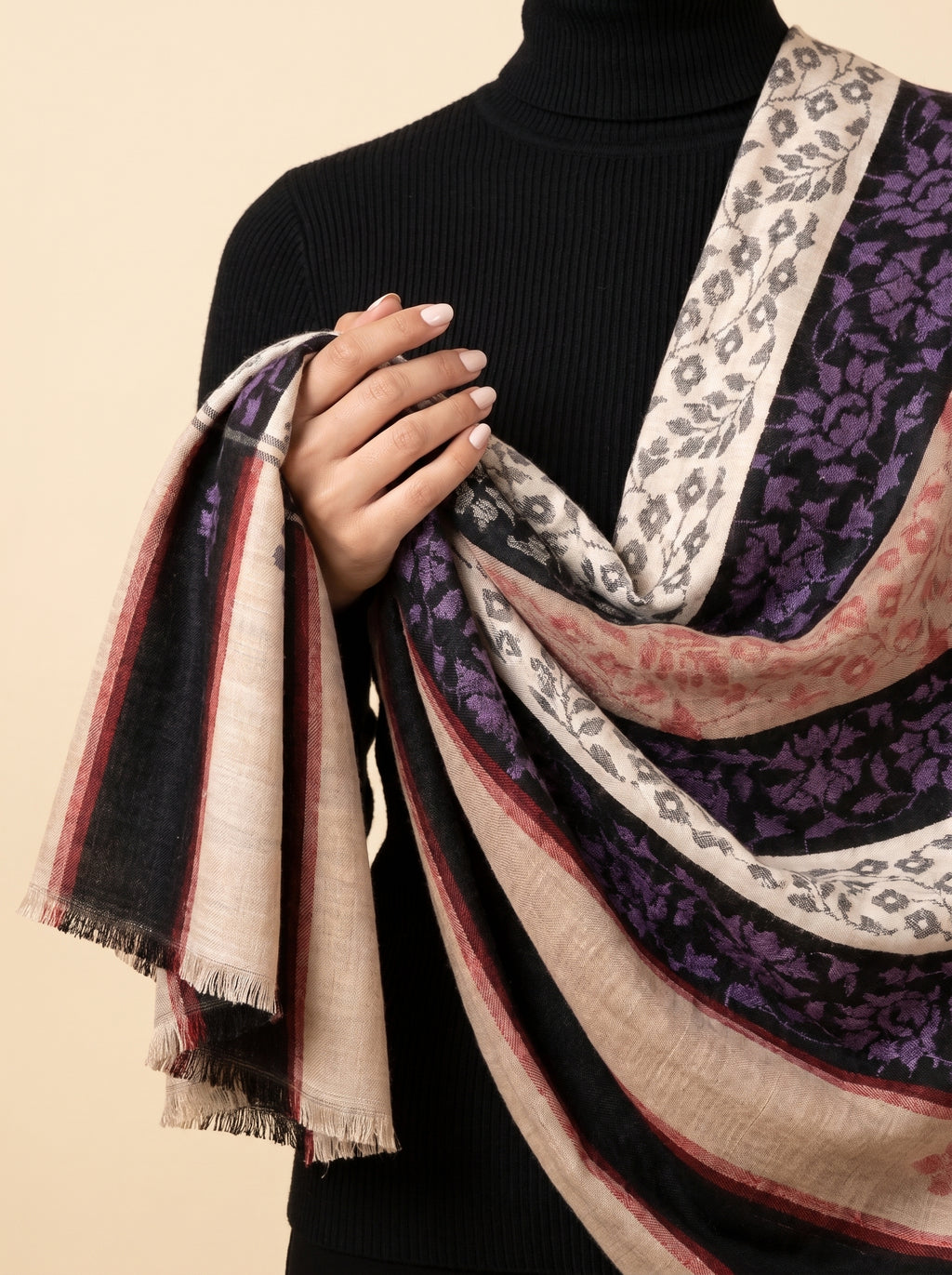 A close-up of a person wearing a black ribbed turtleneck sweater and draping a large patterned scarf. The scarf features multiple colors and patterns including purple floral motifs, gray leopard print, and stripes in beige, black, and red.