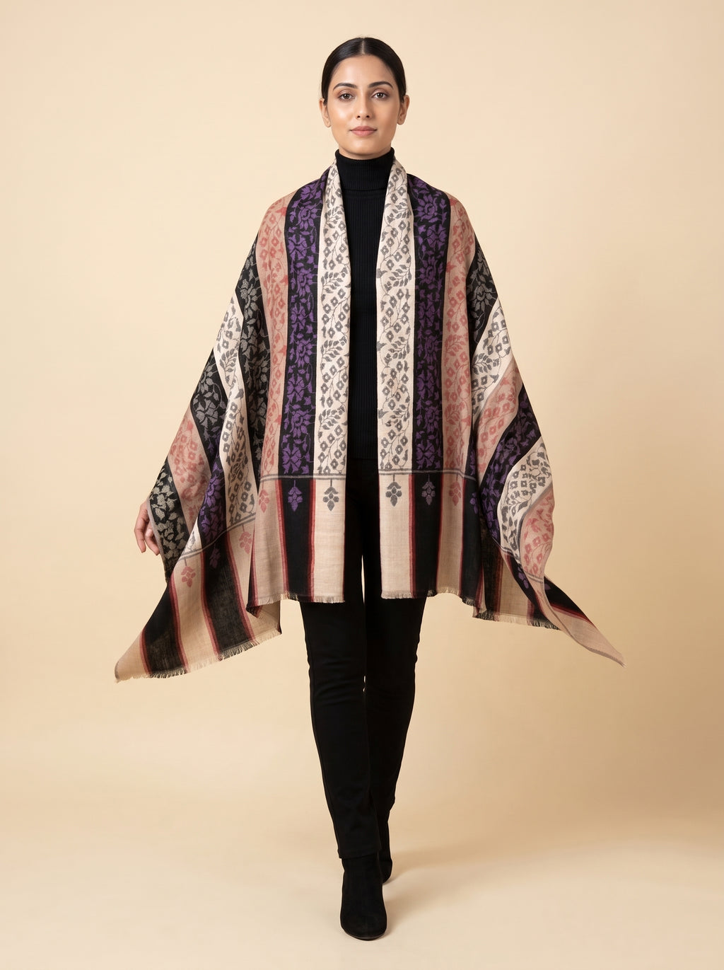 A woman modeling a lightweight open-front shawl with vertical patterned stripes in beige, pink, purple, black, and white, worn over a black turtleneck and black pants against a solid beige studio background.