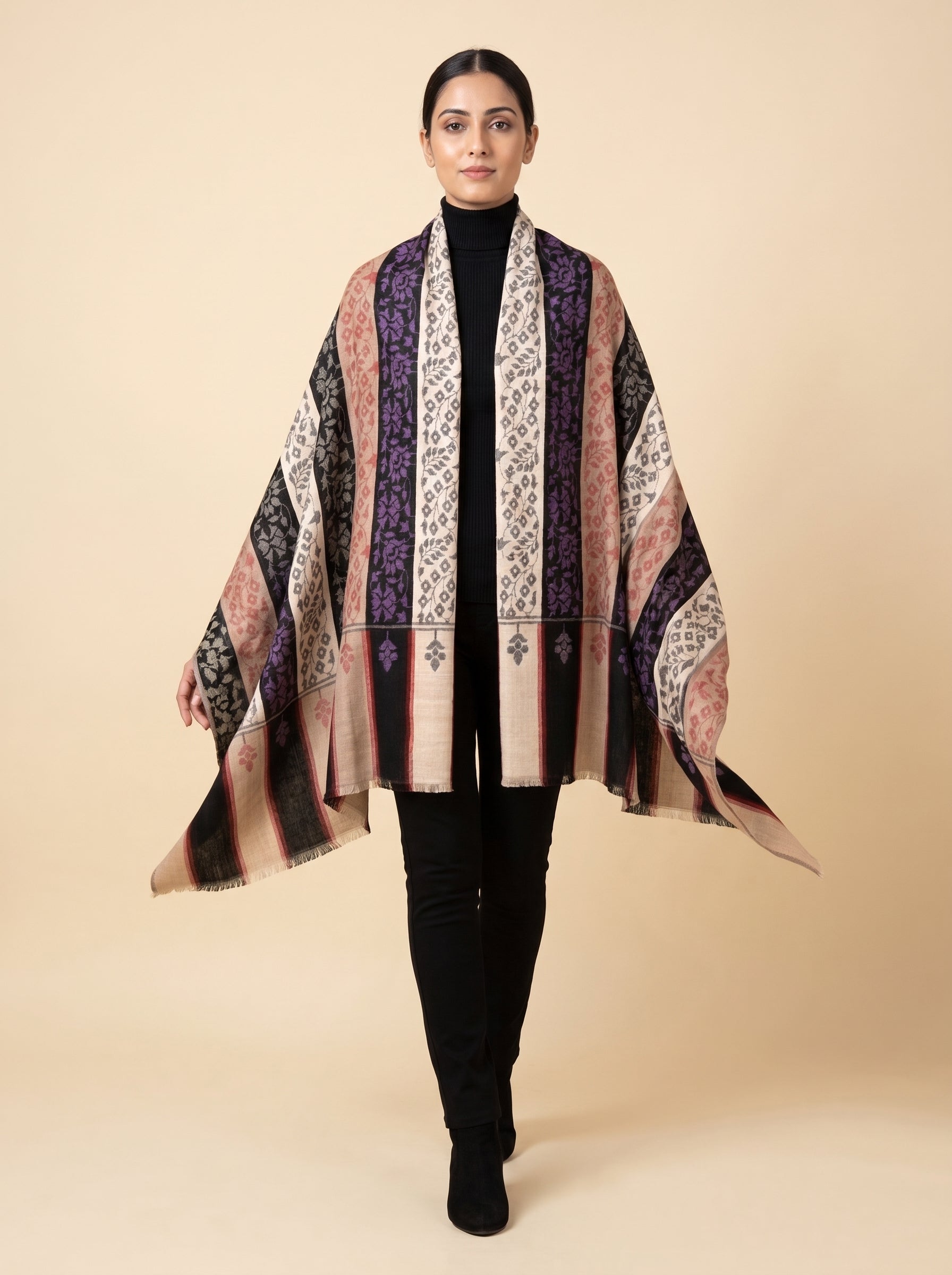 A woman modeling a lightweight open-front shawl with vertical patterned stripes in beige, pink, purple, black, and white, worn over a black turtleneck and black pants against a solid beige studio background.