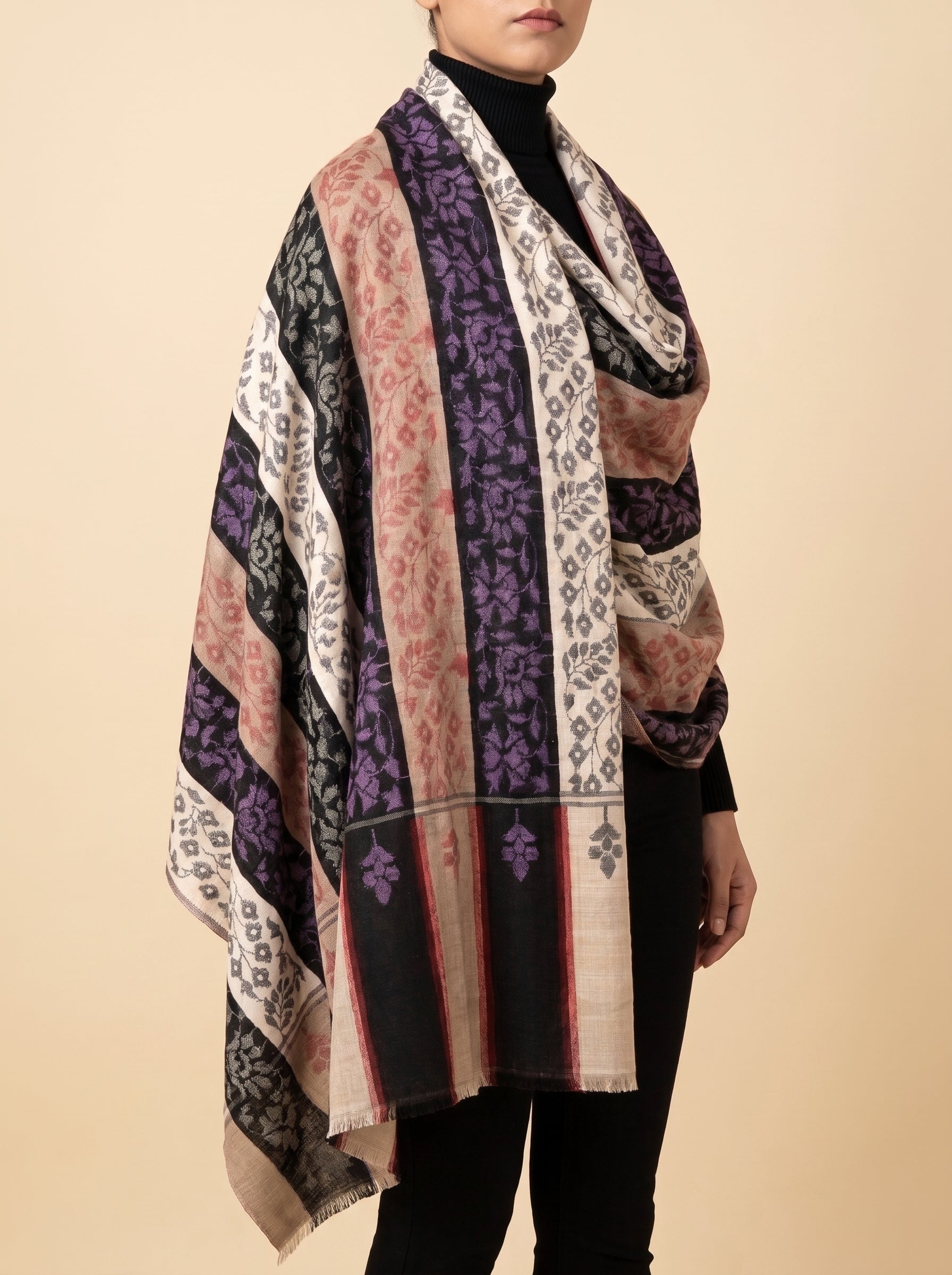 A model wearing a multi-colored patchwork scarf with purple, black, beige, and pink patterns, styled over a black turtleneck against a solid beige background.